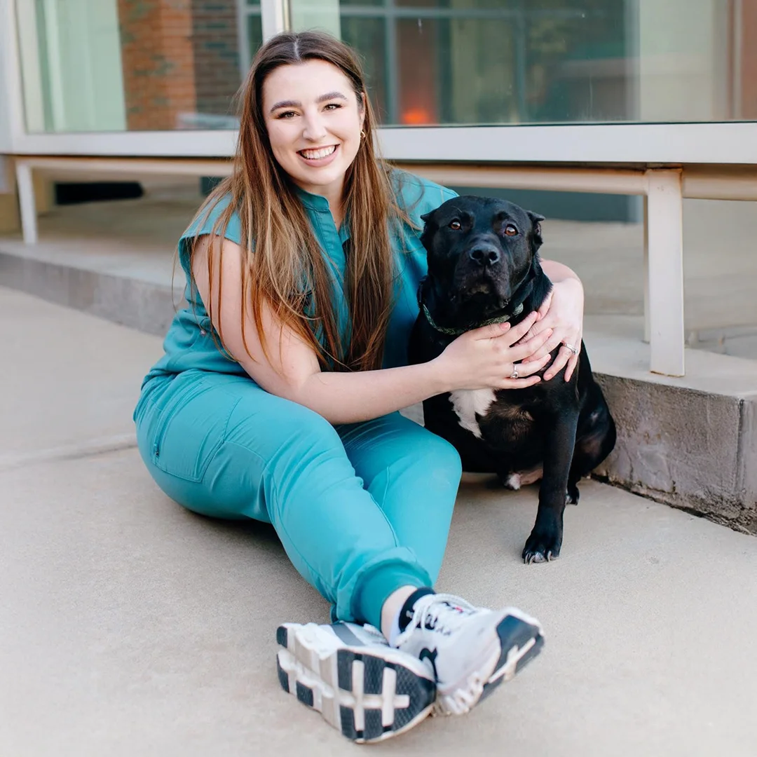 Veterinarian Dr. Ellen Tredway sitting on the sidewalk and smiling while hugging a black dog in front of Animal Medical and Surgical Hospital.
